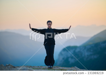Woman practicing Tai Chi in traditional black outfit on mountain at sunrise, arms outstretched in meditation pose focusing on balance, energy, and mindfulness in nature. Woman practicing Tai Chi in traditional black outfit on mountain at sunrise, arms outstretched in meditation pose focusing on balance, energy, and mindfulness in nature. 131069219