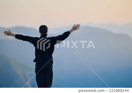 Woman practicing Tai Chi in traditional black outfit on mountain at sunrise, arms outstretched in meditation pose focusing on balance, energy, and mindfulness in nature. 131069244