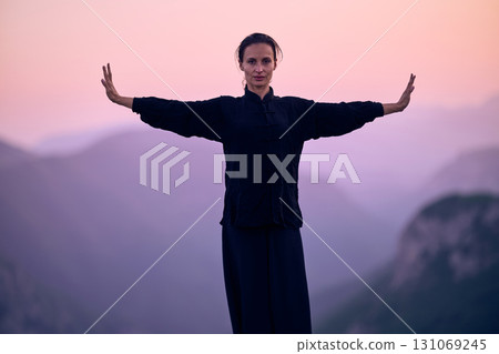 Woman practicing Tai Chi in traditional black outfit on mountain at sunrise, arms outstretched in meditation pose focusing on balance, energy, and mindfulness in nature. 131069245