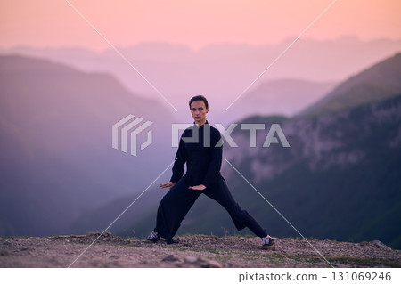 Woman practicing Tai Chi in traditional black outfit on mountain at sunrise, arms outstretched in meditation pose focusing on balance, energy, and mindfulness in nature. 131069246