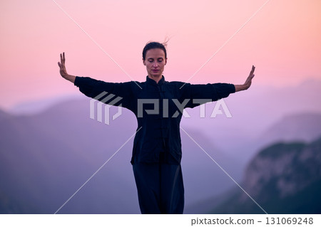 Woman practicing Tai Chi in traditional black outfit on mountain at sunrise, arms outstretched in meditation pose focusing on balance, energy, and mindfulness in nature. 131069248
