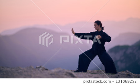 Woman practicing Tai Chi in traditional black outfit on mountain at sunrise, arms outstretched in meditation pose focusing on balance, energy, and mindfulness in nature. 131069252