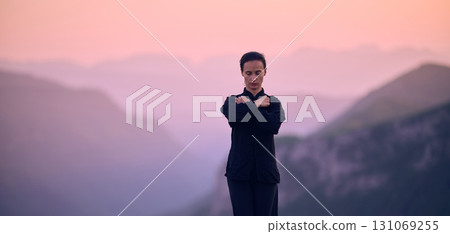 Woman practicing Tai Chi in traditional black outfit on mountain at sunrise, arms outstretched in meditation pose focusing on balance, energy, and mindfulness in nature. 131069255