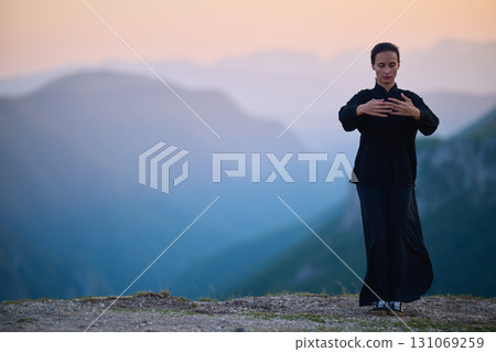 Woman practicing Tai Chi in traditional black outfit on mountain at sunrise, arms outstretched in meditation pose focusing on balance, energy, and mindfulness in nature. 131069259