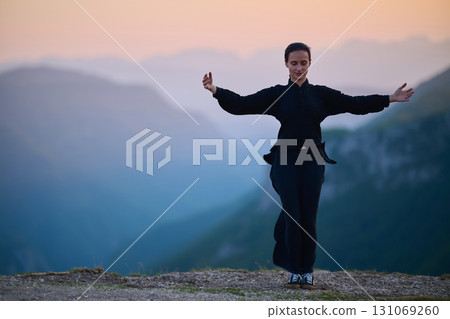 Woman practicing Tai Chi in traditional black outfit on mountain at sunrise, arms outstretched in meditation pose focusing on balance, energy, and mindfulness in nature. Woman practicing Tai Chi in traditional black outfit on mountain at sunrise, arms outstretched in meditation pose focusing on balance, energy, and mindfulness in nature. 131069260