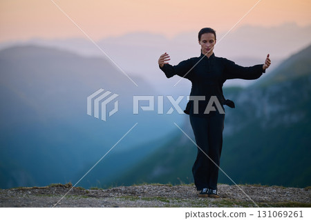 Woman practicing Tai Chi in traditional black outfit on mountain at sunrise, arms outstretched in meditation pose focusing on balance, energy, and mindfulness in nature. Woman practicing Tai Chi in traditional black outfit on mountain at sunrise, arms outstretched in meditation pose focusing on balance, energy, and mindfulness in nature. 131069261