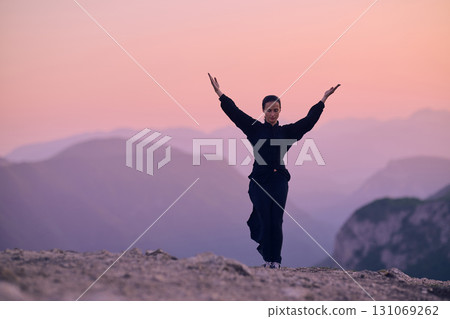Woman practicing Tai Chi in traditional black outfit on mountain at sunrise, arms outstretched in meditation pose focusing on balance, energy, and mindfulness in nature. 131069262