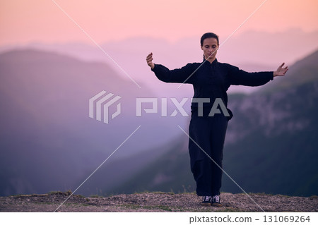 Woman practicing Tai Chi in traditional black outfit on mountain at sunrise, arms outstretched in meditation pose focusing on balance, energy, and mindfulness in nature. Woman practicing Tai Chi in traditional black outfit on mountain at sunrise, arms outstretched in meditation pose focusing on balance, energy, and mindfulness in nature. 131069264