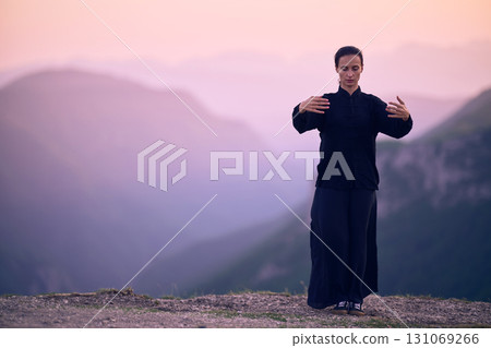 Woman practicing Tai Chi in traditional black outfit on mountain at sunrise, arms outstretched in meditation pose focusing on balance, energy, and mindfulness in nature. 131069266