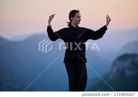 Woman practicing Tai Chi in traditional black outfit on mountain at sunrise, arms outstretched in meditation pose focusing on balance, energy, and mindfulness in nature. 131069267