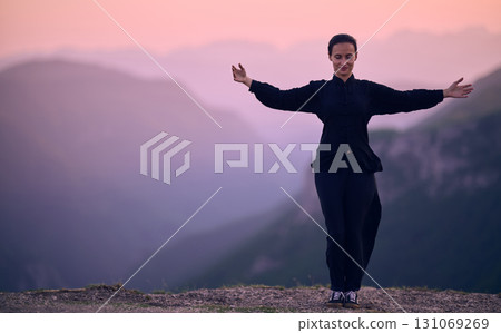 Woman practicing Tai Chi in traditional black outfit on mountain at sunrise, arms outstretched in meditation pose focusing on balance, energy, and mindfulness in nature. 131069269