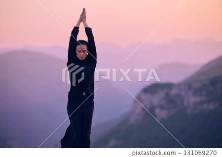Woman practicing Tai Chi in traditional black outfit on mountain at sunrise, arms outstretched in meditation pose focusing on balance, energy, and mindfulness in nature. 131069270