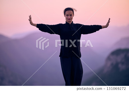Woman practicing Tai Chi in traditional black outfit on mountain at sunrise, arms outstretched in meditation pose focusing on balance, energy, and mindfulness in nature. Woman practicing Tai Chi in traditional black outfit on mountain at sunrise, arms outstretched in meditation pose focusing on balance, energy, and mindfulness in nature. 131069272