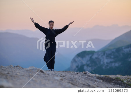 Woman practicing Tai Chi in traditional black outfit on mountain at sunrise, arms outstretched in meditation pose focusing on balance, energy, and mindfulness in nature. Woman practicing Tai Chi in traditional black outfit on mountain at sunrise, arms outstretched in meditation pose focusing on balance, energy, and mindfulness in nature. 131069273