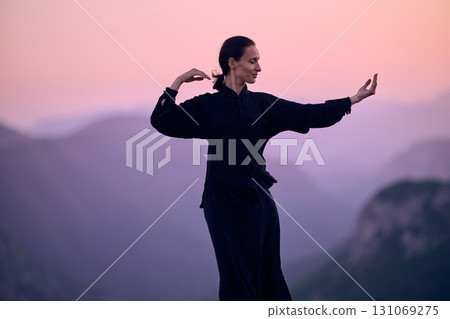 Woman practicing Tai Chi in traditional black outfit on mountain at sunrise, arms outstretched in meditation pose focusing on balance, energy, and mindfulness in nature. 131069275