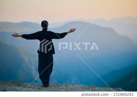 Woman practicing Tai Chi in traditional black outfit on mountain at sunrise, arms outstretched in meditation pose focusing on balance, energy, and mindfulness in nature. 131069281
