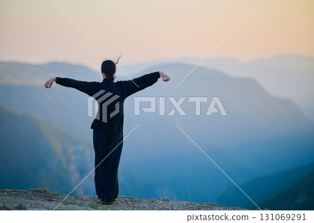 Woman practicing Tai Chi in traditional black outfit on mountain at sunrise, arms outstretched in meditation pose focusing on balance, energy, and mindfulness in nature. 131069291