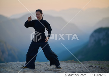 Woman practicing Tai Chi in traditional black outfit on mountain at sunrise, arms outstretched in meditation pose focusing on balance, energy, and mindfulness in nature. 131069292