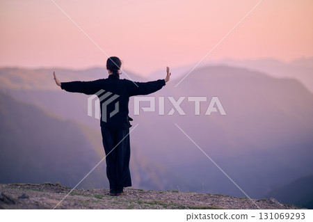 Woman practicing Tai Chi in traditional black outfit on mountain at sunrise, arms outstretched in meditation pose focusing on balance, energy, and mindfulness in nature. Woman practicing Tai Chi in traditional black outfit on mountain at sunrise, arms outstretched in meditation pose focusing on balance, energy, and mindfulness in nature. 131069293