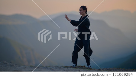 Woman practicing Tai Chi in traditional black outfit on mountain at sunrise, arms outstretched in meditation pose focusing on balance, energy, and mindfulness in nature. Woman practicing Tai Chi in traditional black outfit on mountain at sunrise, arms outstretched in meditation pose focusing on balance, energy, and mindfulness in nature. 131069294