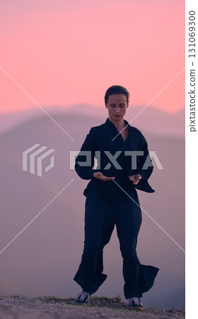 Woman practicing Tai Chi in traditional black outfit on mountain at sunrise, arms outstretched in meditation pose focusing on balance, energy, and mindfulness in nature. 131069300