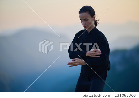 Woman practicing Tai Chi in traditional black outfit on mountain at sunrise, arms outstretched in meditation pose focusing on balance, energy, and mindfulness in nature. 131069306
