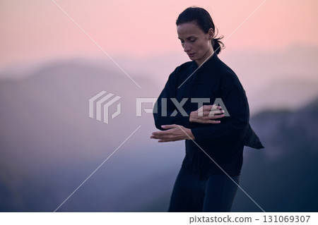 Woman practicing Tai Chi in traditional black outfit on mountain at sunrise, arms outstretched in meditation pose focusing on balance, energy, and mindfulness in nature. 131069307