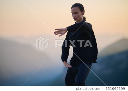 Woman practicing Tai Chi in traditional black outfit on mountain at sunrise, arms outstretched in meditation pose focusing on balance, energy, and mindfulness in nature. 131069309