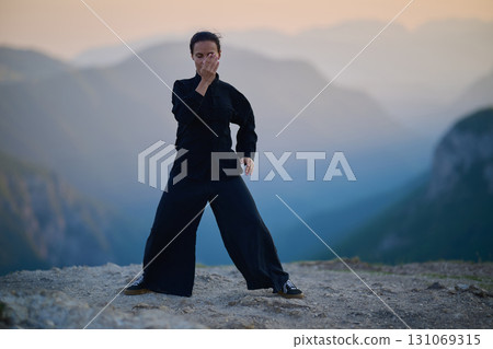 Woman practicing Tai Chi in traditional black outfit on mountain at sunrise, arms outstretched in meditation pose focusing on balance, energy, and mindfulness in nature. 131069315