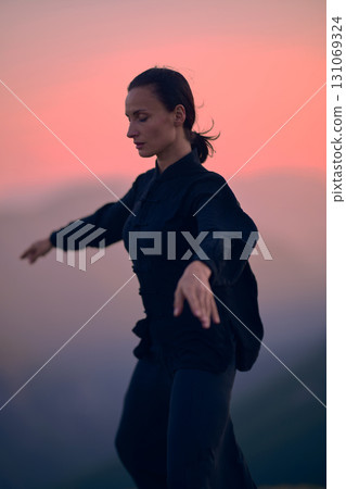 Woman practicing Tai Chi in traditional black outfit on mountain at sunrise, arms outstretched in meditation pose focusing on balance, energy, and mindfulness in nature. Woman practicing Tai Chi in traditional black outfit on mountain at sunrise, arms outstretched in meditation pose focusing on balance, energy, and mindfulness in nature. 131069324