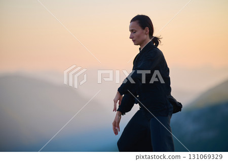 Woman practicing Tai Chi in traditional black outfit on mountain at sunrise, arms outstretched in meditation pose focusing on balance, energy, and mindfulness in nature. 131069329