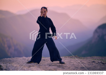 Woman practicing Tai Chi in traditional black outfit on mountain at sunrise, arms outstretched in meditation pose focusing on balance, energy, and mindfulness in nature. Woman practicing Tai Chi in traditional black outfit on mountain at sunrise, arms outstretched in meditation pose focusing on balance, energy, and mindfulness in nature. 131069337