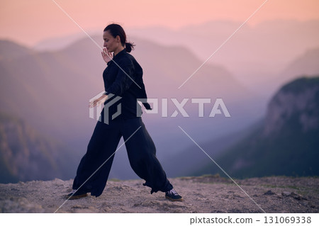Woman practicing Tai Chi in traditional black outfit on mountain at sunrise, arms outstretched in meditation pose focusing on balance, energy, and mindfulness in nature. Woman practicing Tai Chi in traditional black outfit on mountain at sunrise, arms outstretched in meditation pose focusing on balance, energy, and mindfulness in nature. 131069338