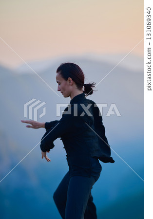 Woman practicing Tai Chi in traditional black outfit on mountain at sunrise, arms outstretched in meditation pose focusing on balance, energy, and mindfulness in nature. 131069340