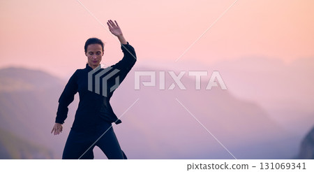Woman practicing Tai Chi in traditional black outfit on mountain at sunrise, arms outstretched in meditation pose focusing on balance, energy, and mindfulness in nature. 131069341