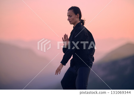 Woman practicing Tai Chi in traditional black outfit on mountain at sunrise, arms outstretched in meditation pose focusing on balance, energy, and mindfulness in nature. Woman practicing Tai Chi in traditional black outfit on mountain at sunrise, arms outstretched in meditation pose focusing on balance, energy, and mindfulness in nature. 131069344