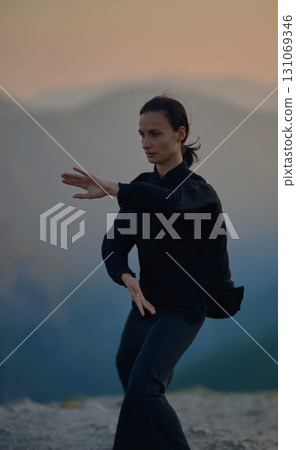 Woman practicing Tai Chi in traditional black outfit on mountain at sunrise, arms outstretched in meditation pose focusing on balance, energy, and mindfulness in nature. Woman practicing Tai Chi in traditional black outfit on mountain at sunrise, arms outstretched in meditation pose focusing on balance, energy, and mindfulness in nature. 131069346