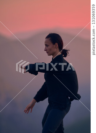Woman practicing Tai Chi in traditional black outfit on mountain at sunrise, arms outstretched in meditation pose focusing on balance, energy, and mindfulness in nature. 131069359