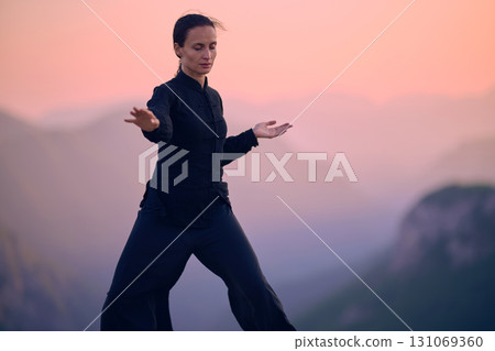 Woman practicing Tai Chi in traditional black outfit on mountain at sunrise, arms outstretched in meditation pose focusing on balance, energy, and mindfulness in nature. 131069360
