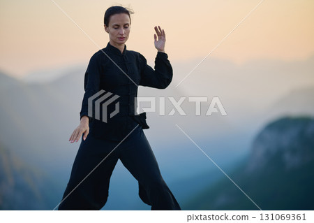 Woman practicing Tai Chi in traditional black outfit on mountain at sunrise, arms outstretched in meditation pose focusing on balance, energy, and mindfulness in nature. Woman practicing Tai Chi in traditional black outfit on mountain at sunrise, arms outstretched in meditation pose focusing on balance, energy, and mindfulness in nature. 131069361