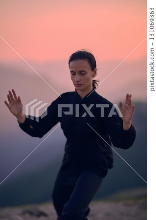 Woman practicing Tai Chi in traditional black outfit on mountain at sunrise, arms outstretched in meditation pose focusing on balance, energy, and mindfulness in nature. Woman practicing Tai Chi in traditional black outfit on mountain at sunrise, arms outstretched in meditation pose focusing on balance, energy, and mindfulness in nature. 131069363