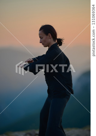 Woman practicing Tai Chi in traditional black outfit on mountain at sunrise, arms outstretched in meditation pose focusing on balance, energy, and mindfulness in nature. 131069364