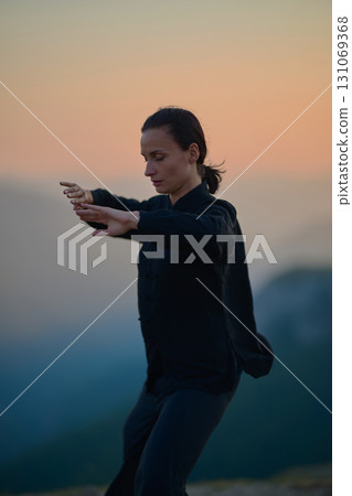 Woman practicing Tai Chi in traditional black outfit on mountain at sunrise, arms outstretched in meditation pose focusing on balance, energy, and mindfulness in nature. 131069368