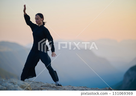 Woman practicing Tai Chi in traditional black outfit on mountain at sunrise, arms outstretched in meditation pose focusing on balance, energy, and mindfulness in nature. 131069369
