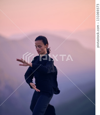 Woman practicing Tai Chi in traditional black outfit on mountain at sunrise, arms outstretched in meditation pose focusing on balance, energy, and mindfulness in nature. 131069373