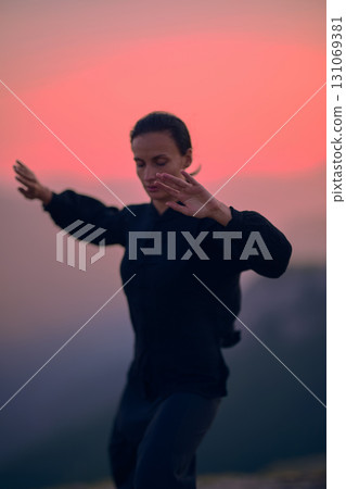 Woman practicing Tai Chi in traditional black outfit on mountain at sunrise, arms outstretched in meditation pose focusing on balance, energy, and mindfulness in nature. 131069381