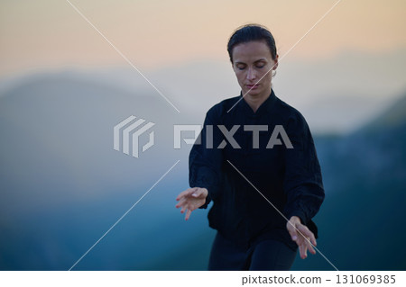 Woman practicing Tai Chi in traditional black outfit on mountain at sunrise, arms outstretched in meditation pose focusing on balance, energy, and mindfulness in nature. 131069385