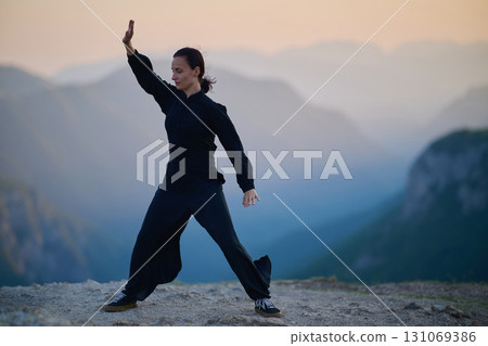 Woman practicing Tai Chi in traditional black outfit on mountain at sunrise, arms outstretched in meditation pose focusing on balance, energy, and mindfulness in nature. 131069386