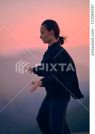 Woman practicing Tai Chi in traditional black outfit on mountain at sunrise, arms outstretched in meditation pose focusing on balance, energy, and mindfulness in nature. Woman practicing Tai Chi in traditional black outfit on mountain at sunrise, arms outstretched in meditation pose focusing on balance, energy, and mindfulness in nature. 131069387