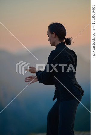 Woman practicing Tai Chi in traditional black outfit on mountain at sunrise, arms outstretched in meditation pose focusing on balance, energy, and mindfulness in nature. 131069403